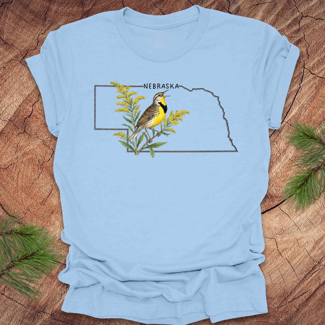 Baby blue t-shirt with the outline of the state of Nebraska and a graphic of a meadowlark and goldenrod inside on a wooden surface