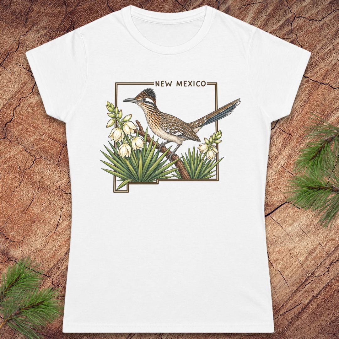 A white short-sleeve crewneck t-shirt featuring a printed graphic of a roadrunner bird perched on a branch with white flowers and green agave plants, framed by the outline of New Mexico with the text 'NEW MEXICO' above. The shirt is displayed flat on a rustic wood background with pine needles in the corners.