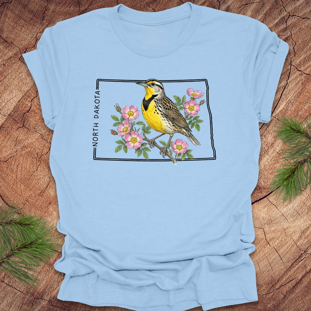 Baby blue t-shirt with a meadowlark and prairie rose flower inside the outline of North Dakota on a wooden surface