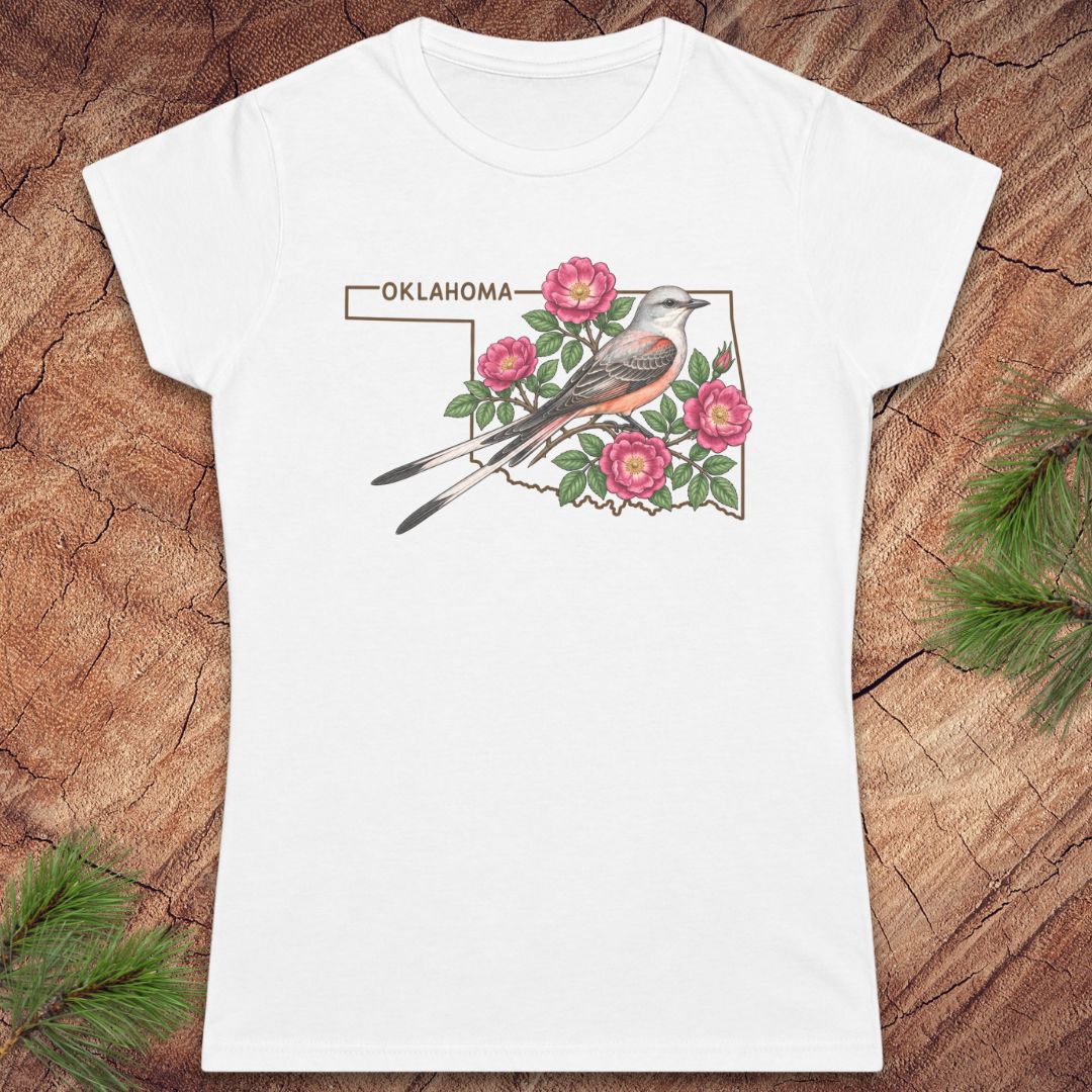 A white short-sleeve crewneck t-shirt featuring a graphic print of an Oklahoma state outline with a bird, pink flowers, and the word 'OKLAHOMA' at the top, displayed flat on a rustic wood background with pine branches.