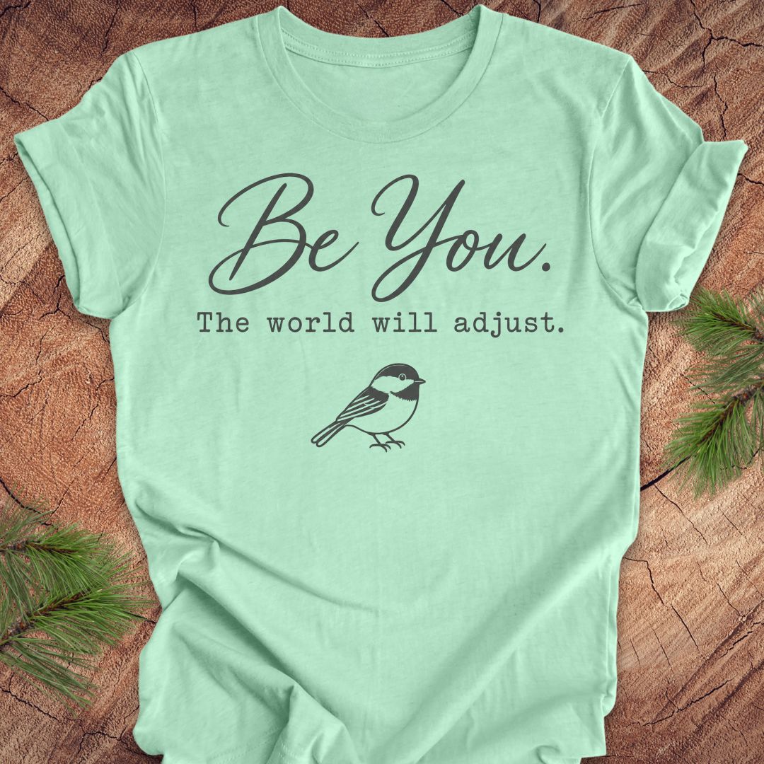A mint green short-sleeve t-shirt with a dark gray graphic print that reads 'Be You. The world will adjust.