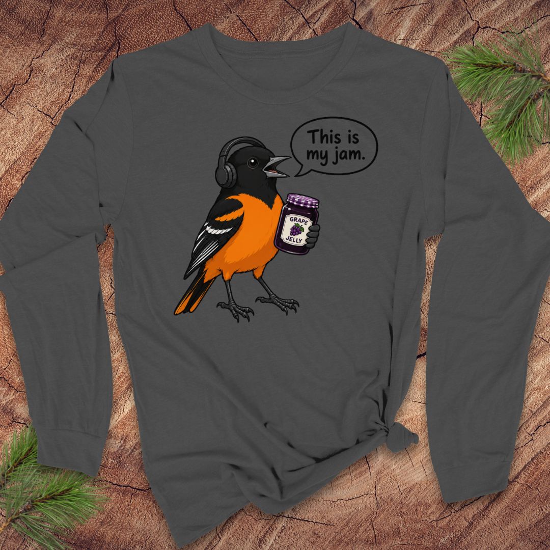 A dark gray long-sleeve t-shirt with a graphic of an oriole bird wearing headphones and holding a jar of jam, with a speech bubble that says 'This is my jam.' The shirt is displayed flat on a rustic wood background with pine branches in the corners.