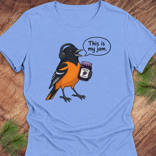 Carolina blue women's relaxed t-shirt with a Baltimore Oriole in headphones holding a grape jelly jar and a 'This is my jam.' speech bubble, laid flat on a rustic wood background with pine branches.