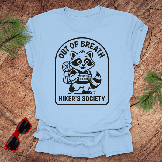 Baby blue t-shirt with a raccoon graphic and text "out of breath hiker's society' on a wooden surface with sunglasses and greenery.