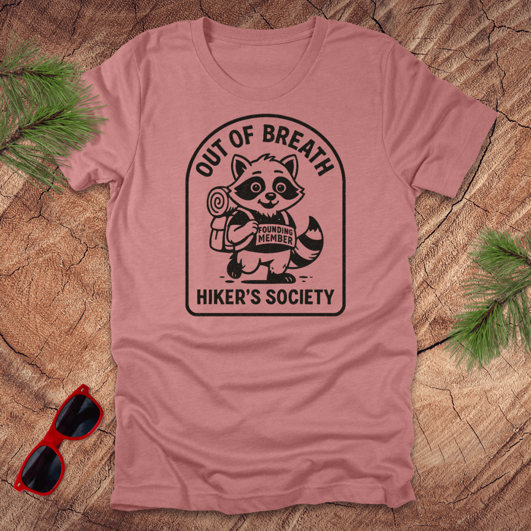 Mauve t-shirt with a raccoon graphic and text "out of breath hiker's society' on a wooden surface with sunglasses and greenery.