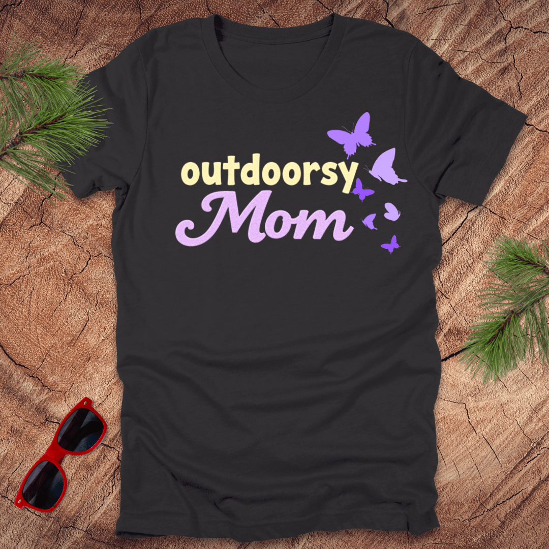 A black t-shirt with the text 'outdoorsy Mom' and butterfly graphics, laid out flat on a wooden background with sunglasses and greenery to the side.