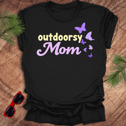 A black t-shirt with the text 'outdoorsy Mom' and butterfly graphics, laid out flat on a wooden background with sunglasses and greenery to the side.