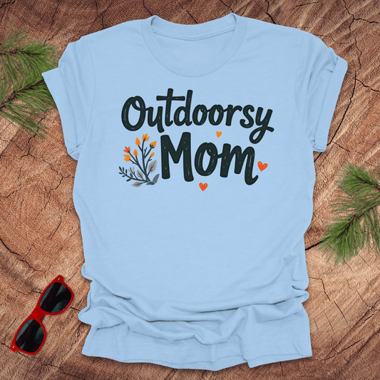 A beige t-shirt with 'Outdoorsy Mom' printed on the front, featuring a floral graphic design with heart accents, displayed on a wooden surface with pine branches and red sunglasses to the side.