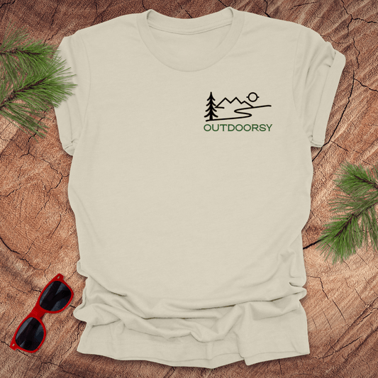 A beige tee displayed flat on a wooden surface, with a minimalist mountain scene and the word 'OUTDOORSY' in pine green on the chest pocket.