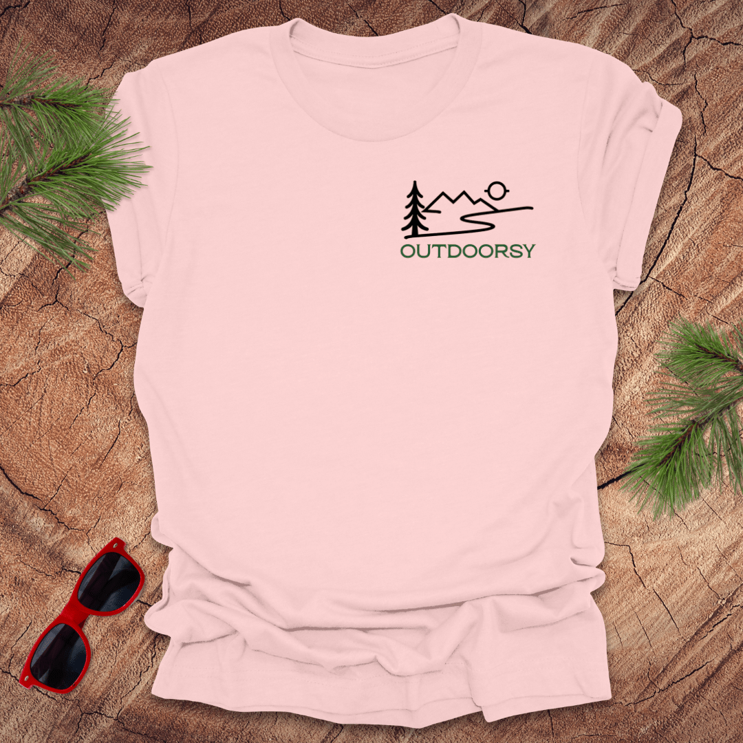 A pink tee displayed flat on a wooden surface, with a minimalist mountain scene and the word 'OUTDOORSY' in pine green on the chest pocket.