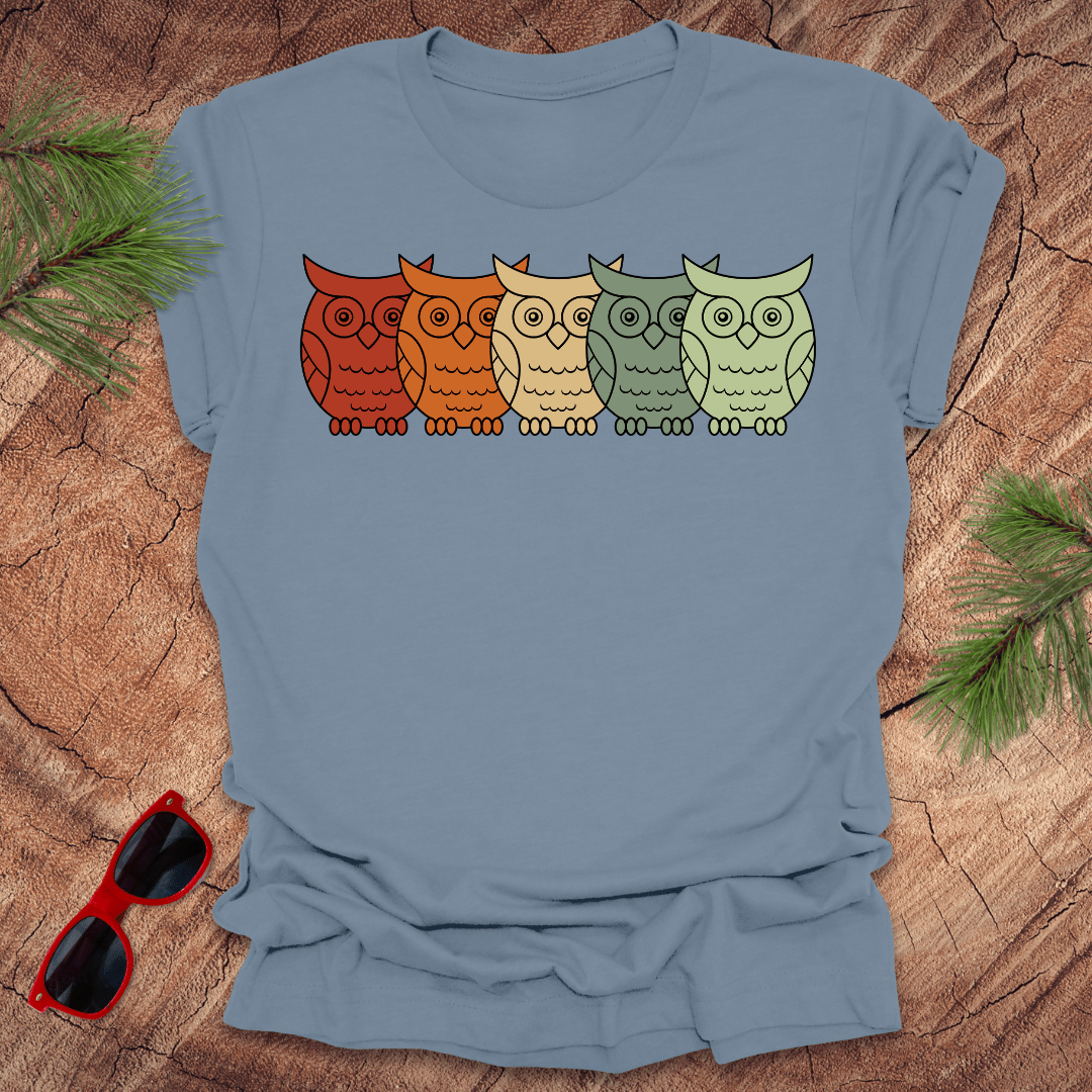 Steel blue tshirt with 5 rainbow colored owls in a line across. On a wood background with pine branches and sunglasses.