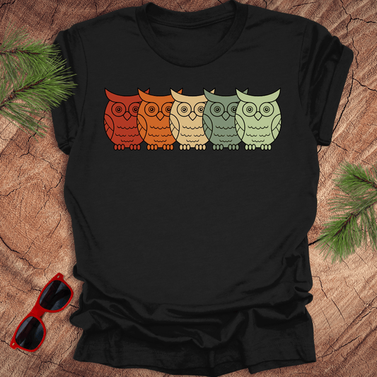 Black tshirt with 5 rainbow colored owls in a line across. On a wood background with pine branches and sunglasses.