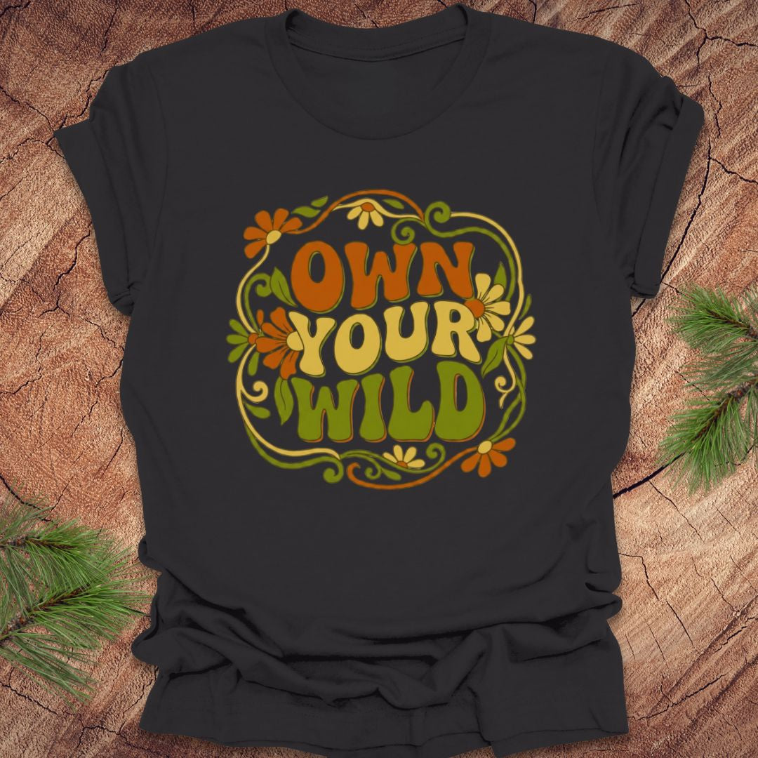 Dark gray t-shirt with 'Own Your Wild' floral design on a wooden surface with greenery.