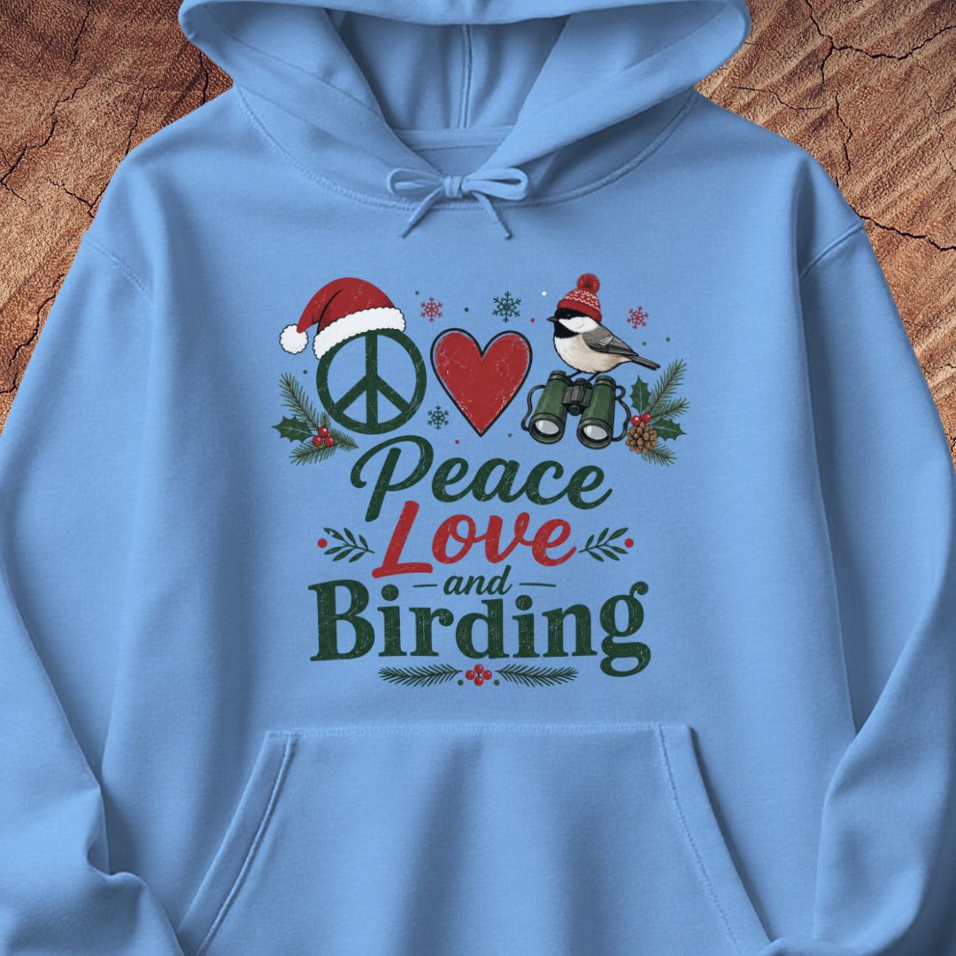 A light blue hoodie laid flat on a rustic wood background, featuring a festive front graphic with a peace sign, red heart, bird, binoculars, pine branches, and the words 'Peace Love and Birding' in mixed dark and red lettering.
