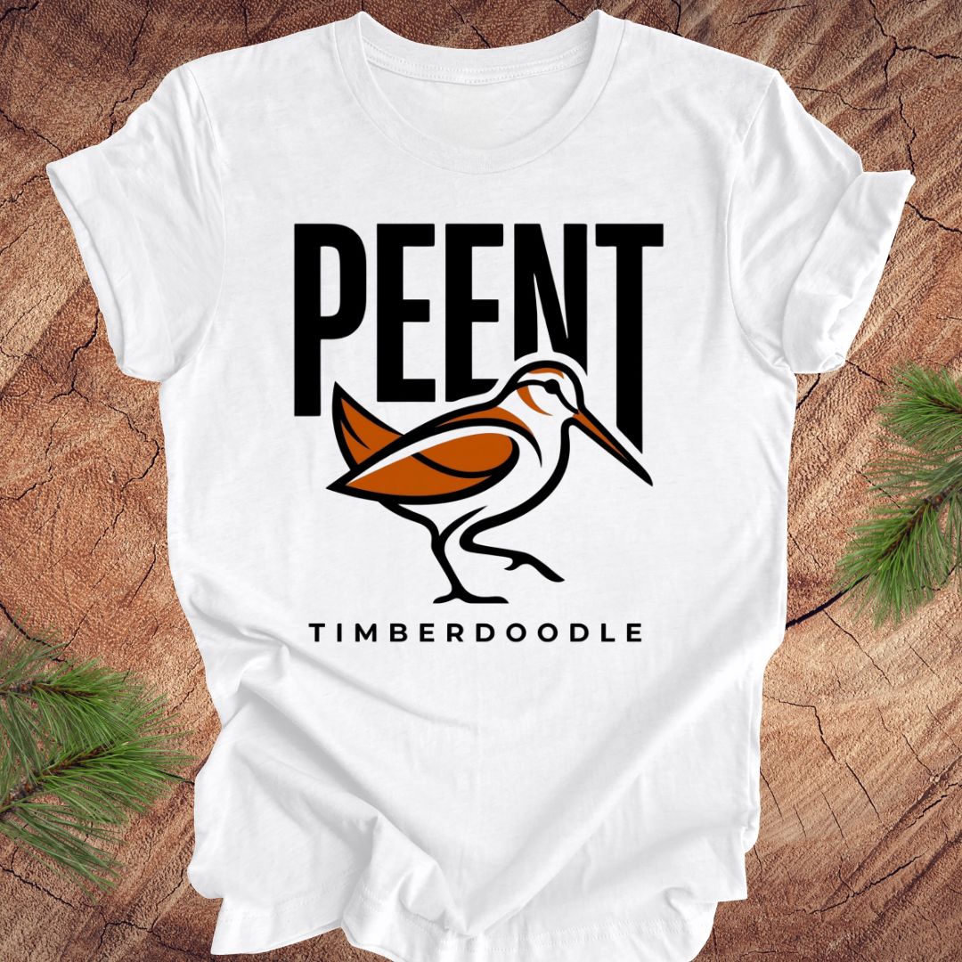 White t-shirt with 'PEENT' and 'TIMBERDOODLE' woodcock design on a wooden surface.
