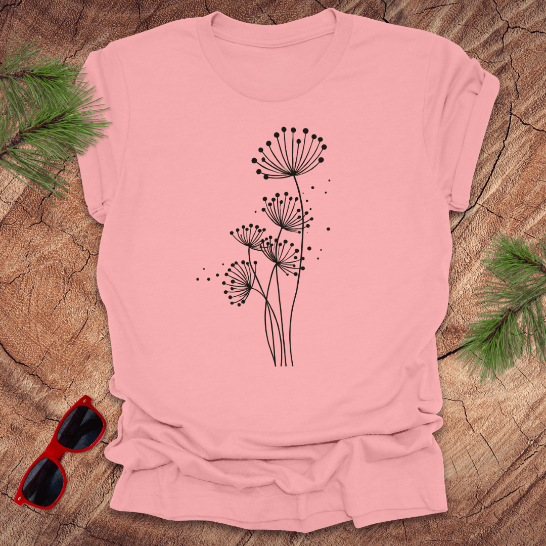 Pink t-shirt with black floral dandelion design on a wooden surface with sunglasses and greenery.