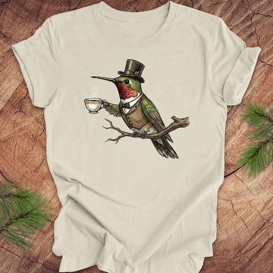 Beige t-shirt with a hummingbird wearing a top hat and holding a cup design on a wooden surface.