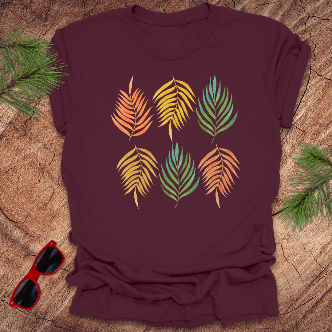A maroon t-shirt with a vibrant leaf pattern in rainbow tones, displayed on a wooden background with foliage accents.