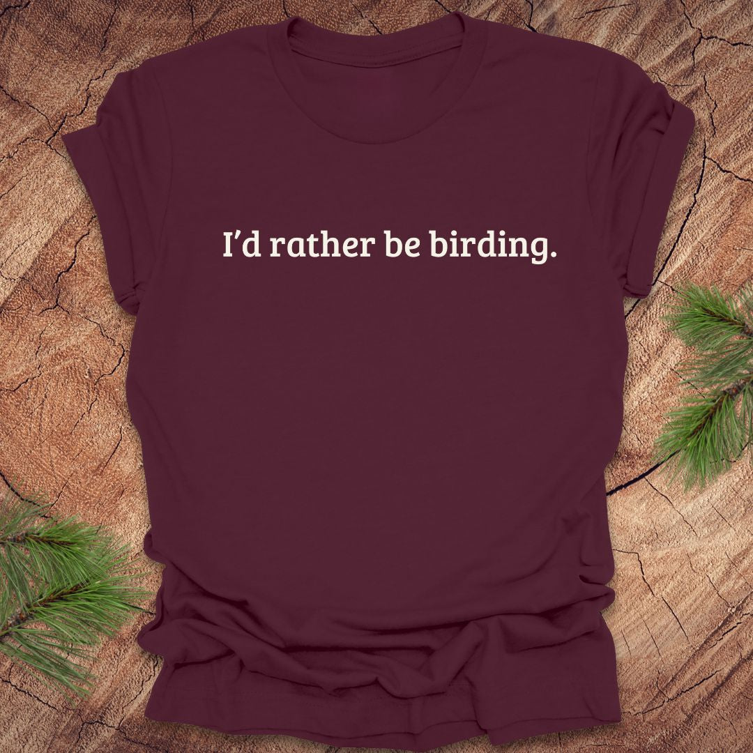 Maroon t-shirt with 'I'd rather be birding.' text on a wooden surface with greenery.