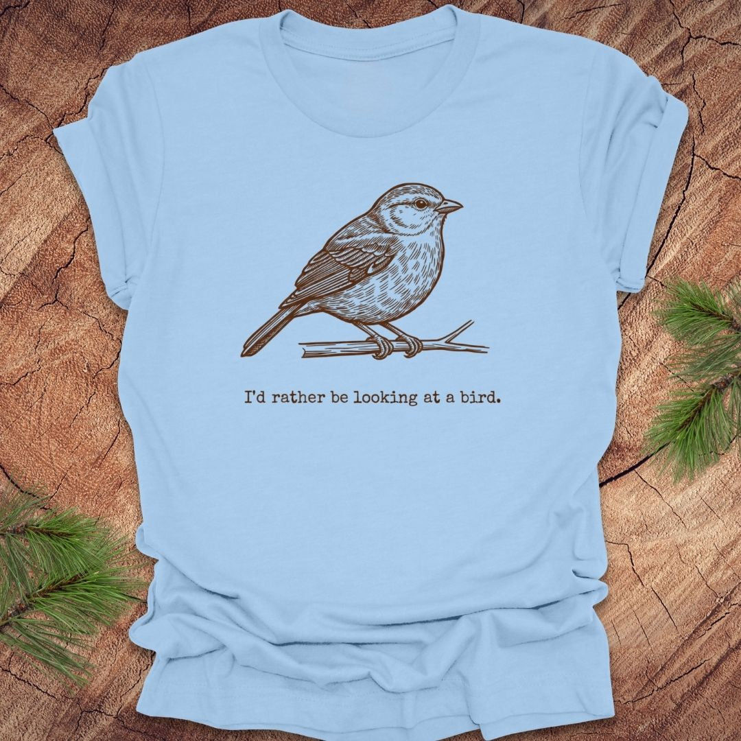 Baby blue t-shirt with a hand drawn brown bird on a branch with text, 'i'd rather be looking at a bird.' on a wooden background.