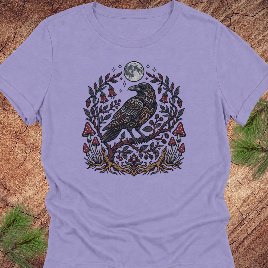A lavender purple t-shirt featuring a detailed graphic of a raven perched on a branch with red mushrooms, green leaves, and a full moon, laid flat on a rustic wood background with pine needles.
