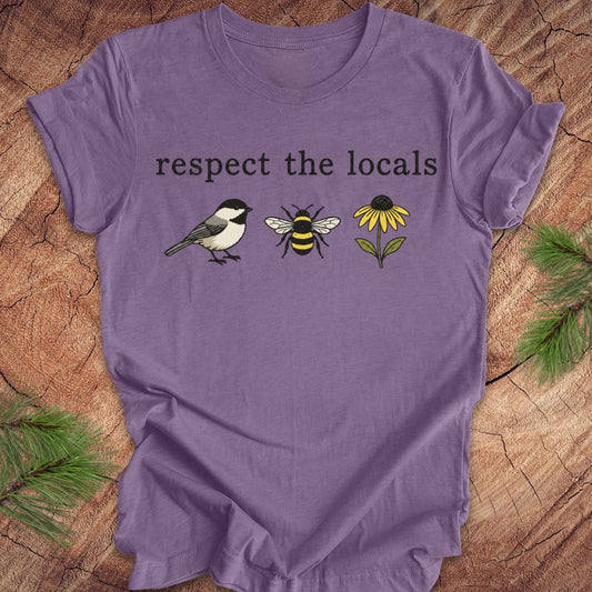 A purple short-sleeve t-shirt laid flat on a rustic wood background with pine branches in the corners. The shirt features the text 'respect the locals' above a graphic of a chickadee, a bumblebee, and a yellow flower.