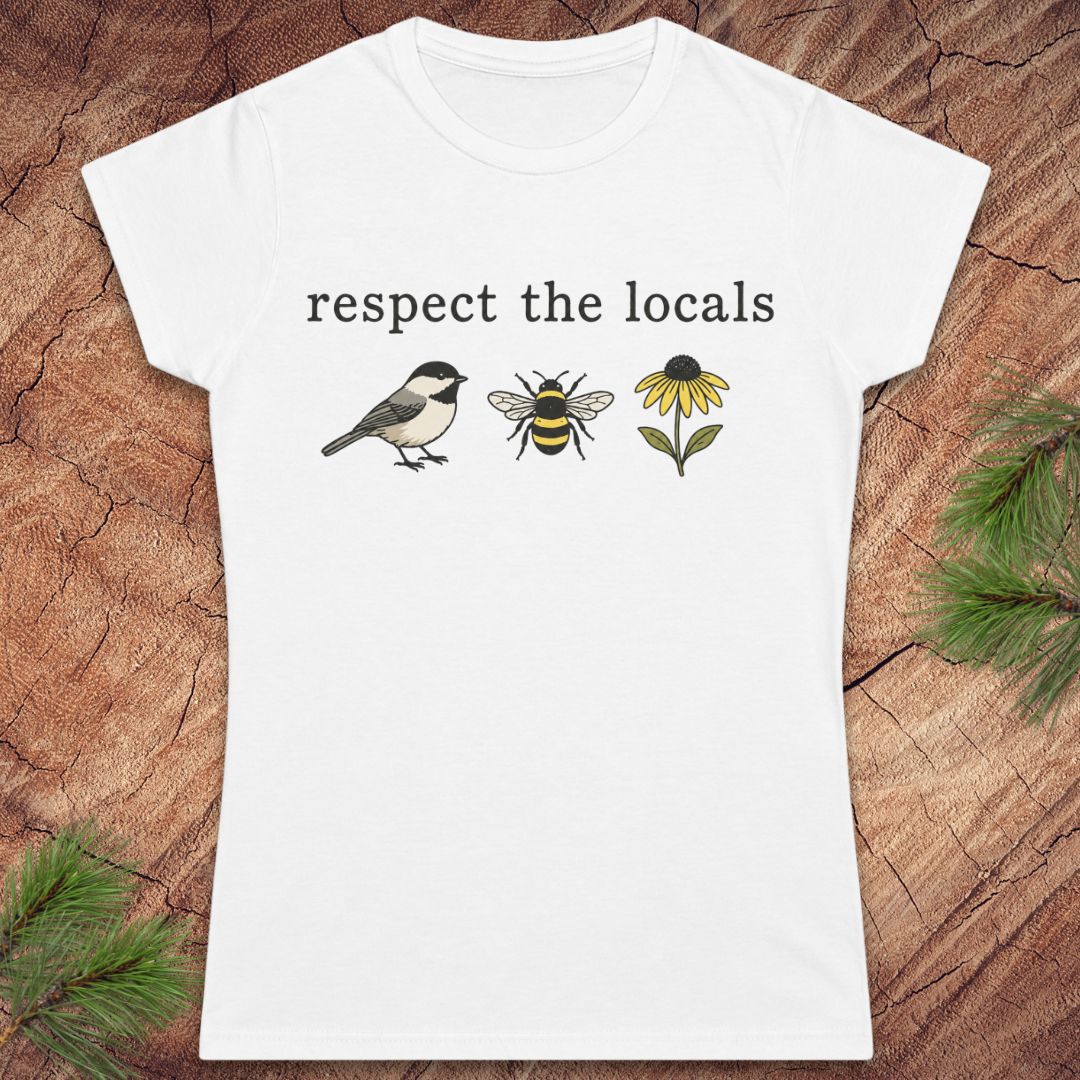 A white short-sleeve crewneck t-shirt with a graphic print on the front reading 'respect the locals' above a chickadee, a bee, and a yellow flower. The shirt is displayed flat on a textured brown wood background with pine needles in the corners.