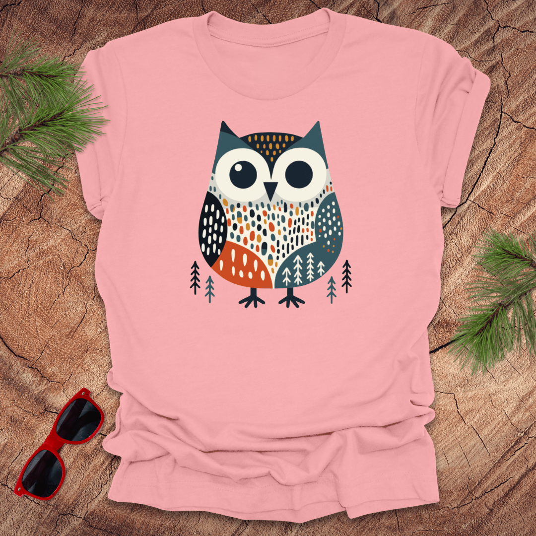 Pink t-shirt with an owl design on a wooden surface with sunglasses and pine branches.