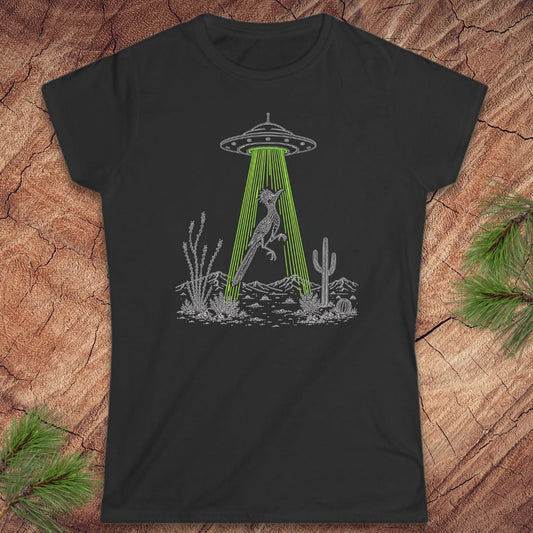 A black short-sleeve crewneck t-shirt laid flat on a textured wood background with pine branches in the corners. The shirt features a black line art graphic of a bird under a UFO with green rays shining down toward a desert landscape.