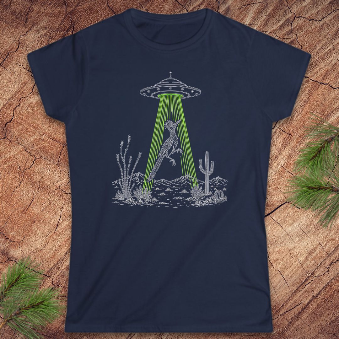 A navy blue short-sleeve crewneck t-shirt laid flat on a textured wood background with pine branches in the corners. The shirt features a black line art graphic of a bird under a UFO with green rays shining down toward a desert landscape.