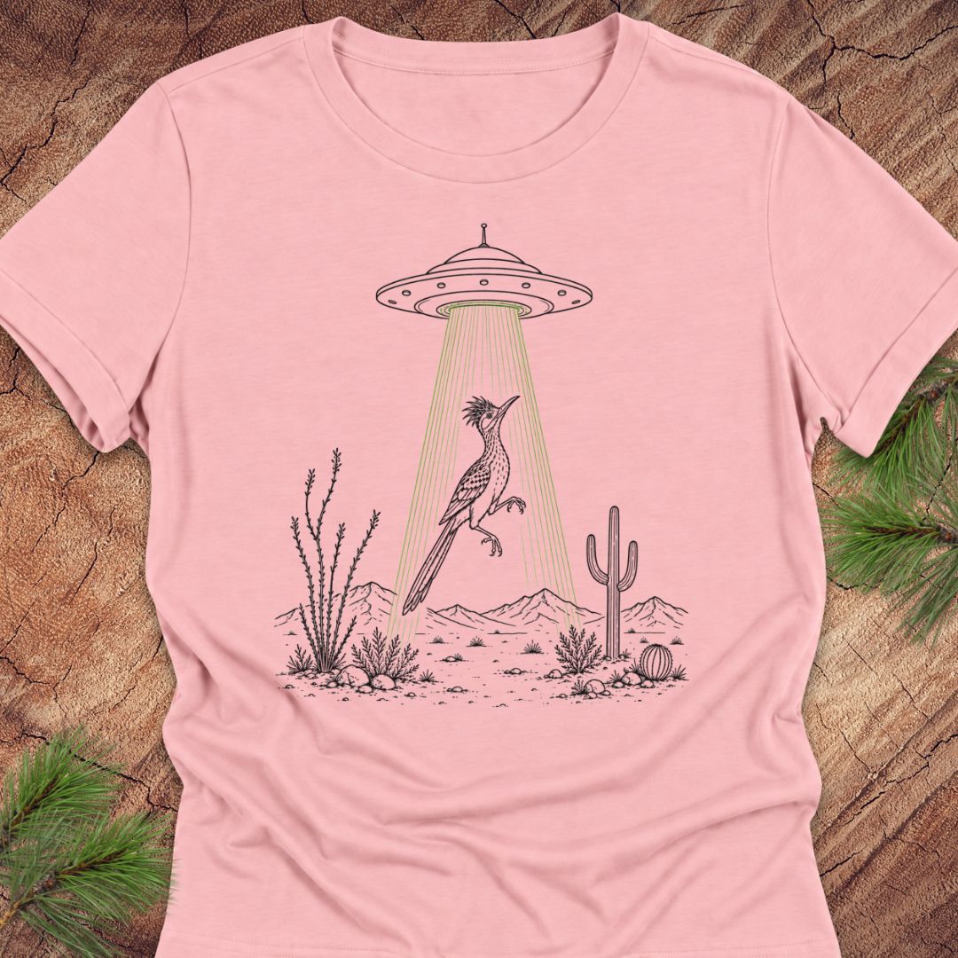 A light pink short-sleeve crewneck t-shirt laid flat on a rustic wood background with pine needles in the corners. The shirt features a graphic print of a desert scene with a cactus, saguaro, and a bird looking up at a UFO.