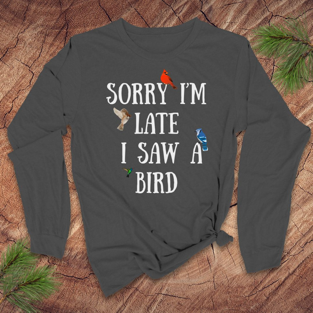 Gray long-sleeve shirt with text 'Sorry I'm late I saw a bird' on a wooden surface with pine branches.