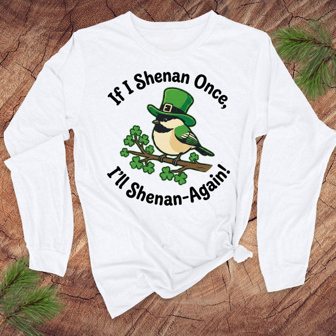 White long-sleeve shirt with a graphic of a bird wearing a green hat and shamrocks, text 'If I Shenan Once, I'll Shenan Again!' on a wooden surface.