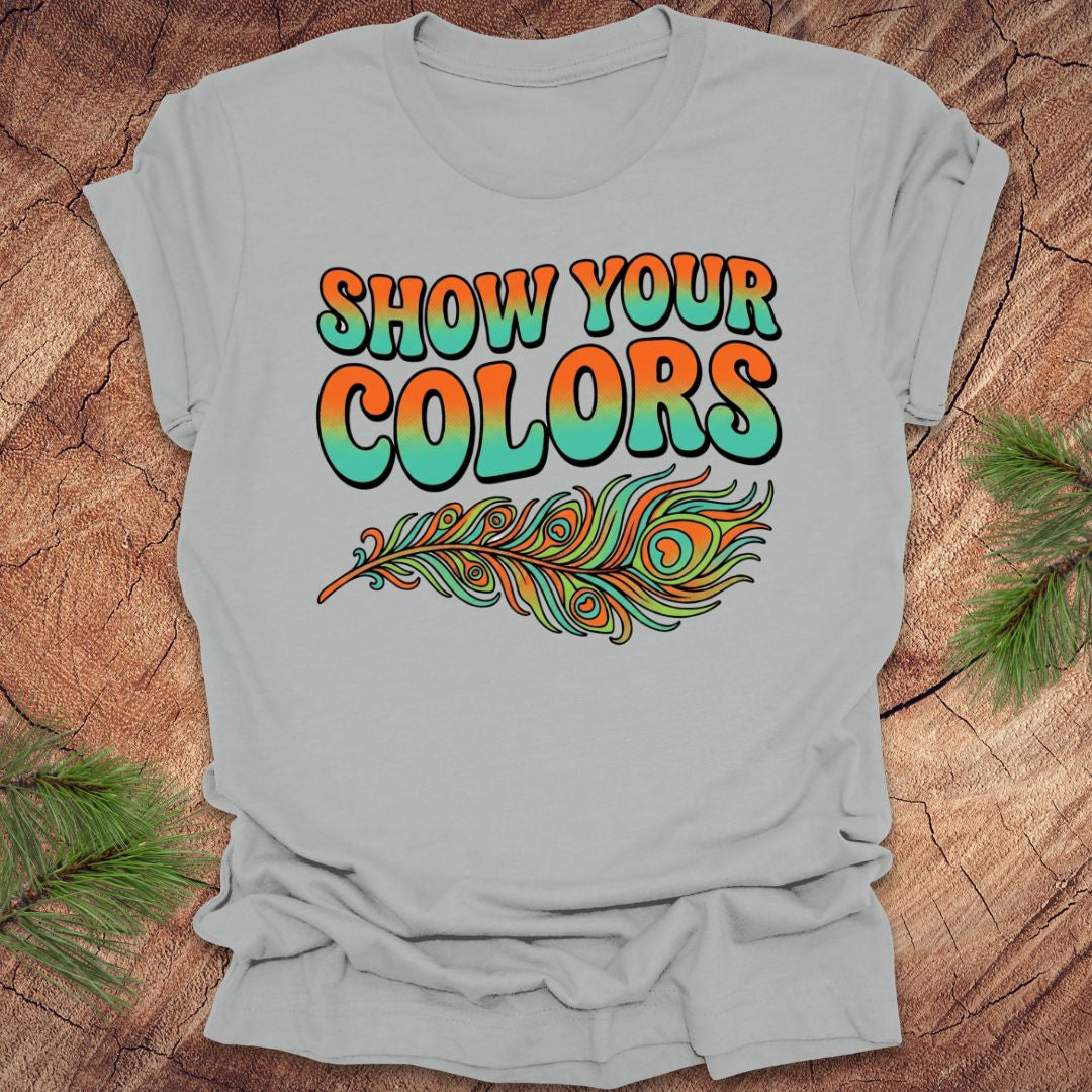 Silver t-shirt with 'Show Your Colors' text and peacock feather design on a wooden surface.