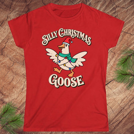 Red women's t-shirt with a goose in a Santa hat and teal scarf, wrapped in colorful Christmas lights under retro Silly Christmas Goose lettering, laid flat on a rustic wood background with pine branches.