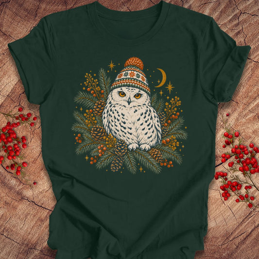 A dark forest green t-shirt laid flat on a rustic wood background, featuring a central graphic of a white owl wearing a colorful knit winter hat, surrounded by pine branches, berries, pinecones, and a crescent moon.