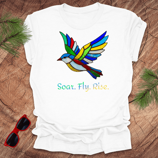 White t-shirt with a colorful bird design and 'Soar. Fly. Rise.' text on a wooden surface with sunglasses and greenery.