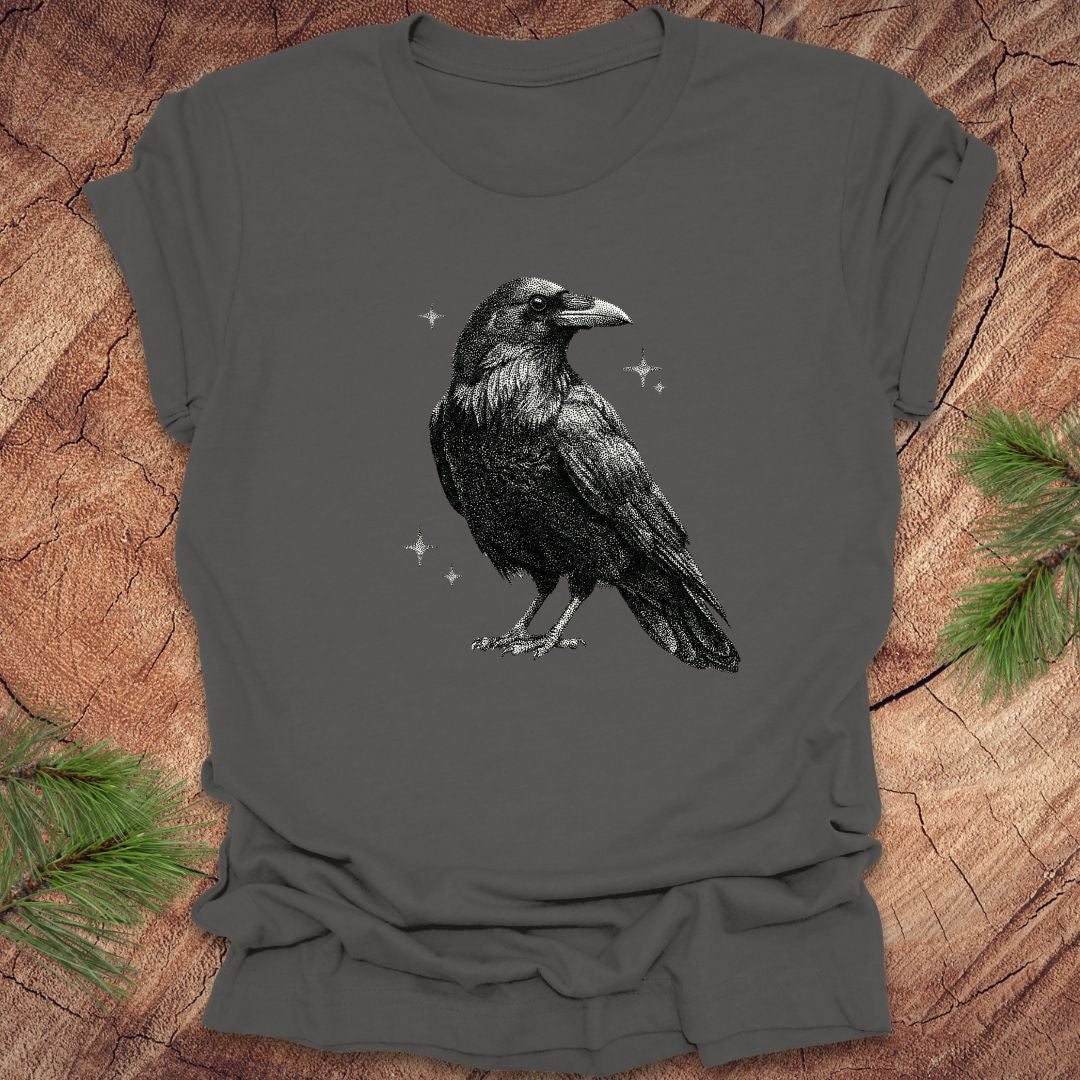Gray t-shirt with a black raven design on a wooden surface with pine branches.