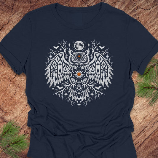 A navy blue short-sleeve t-shirt with a detailed white owl graphic on the chest, laid flat on a rustic wood background with pine branches in the corners.
