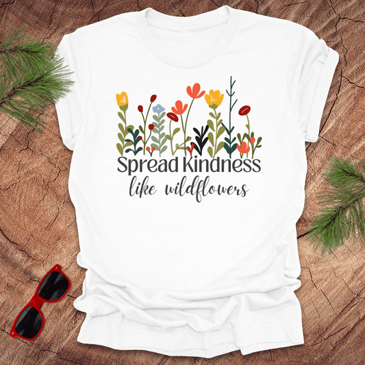 White t-shirt with floral design and 'Spread kindness like wildflowers' text on a wooden surface with sunglasses and greenery.