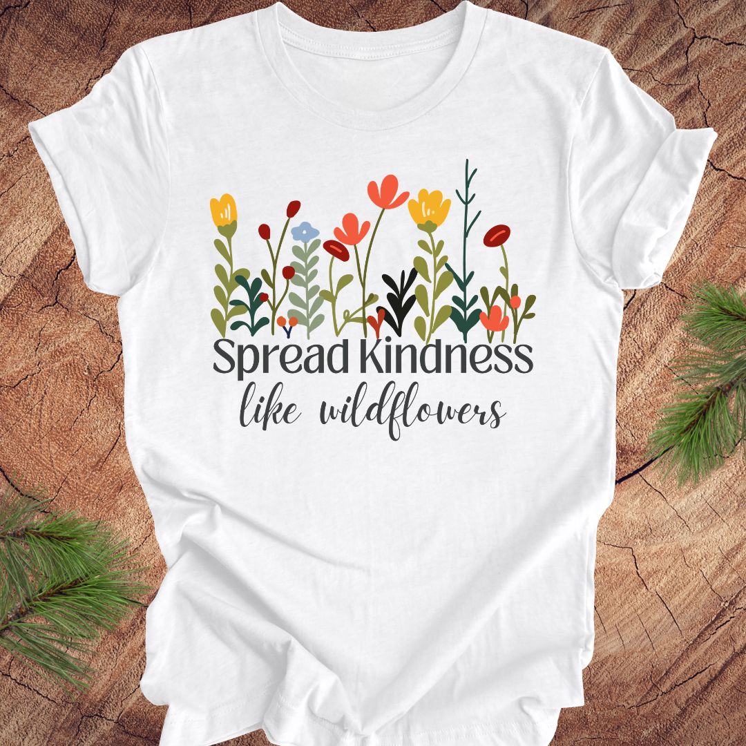 White t-shirt with floral design and 'Spread kindness like wildflowers' text on a wooden surface.