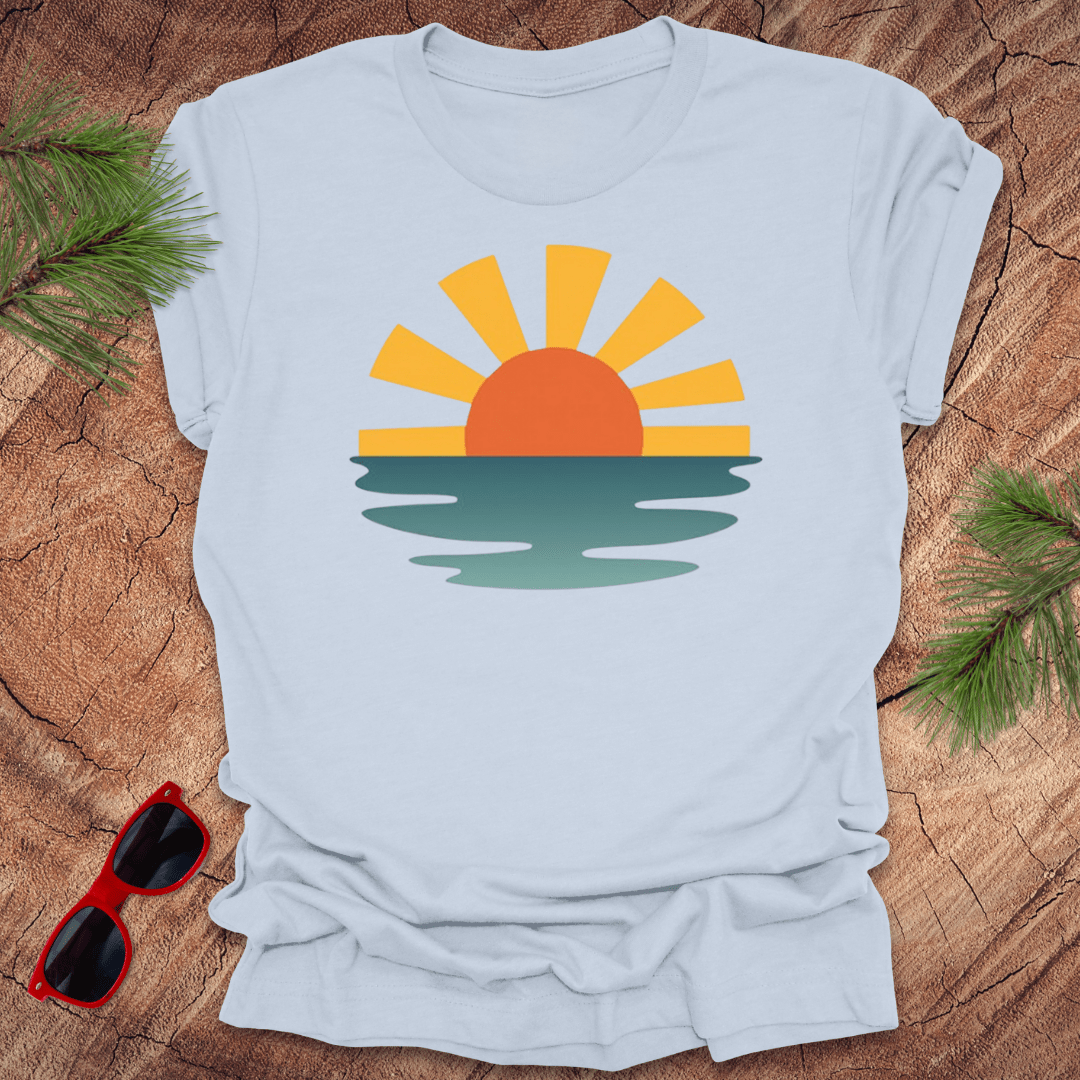 A light blue unisex tee with a graphic print of a sunset over a body of water on the front, displayed on a wooden background with sunglasses and greenery to the side.