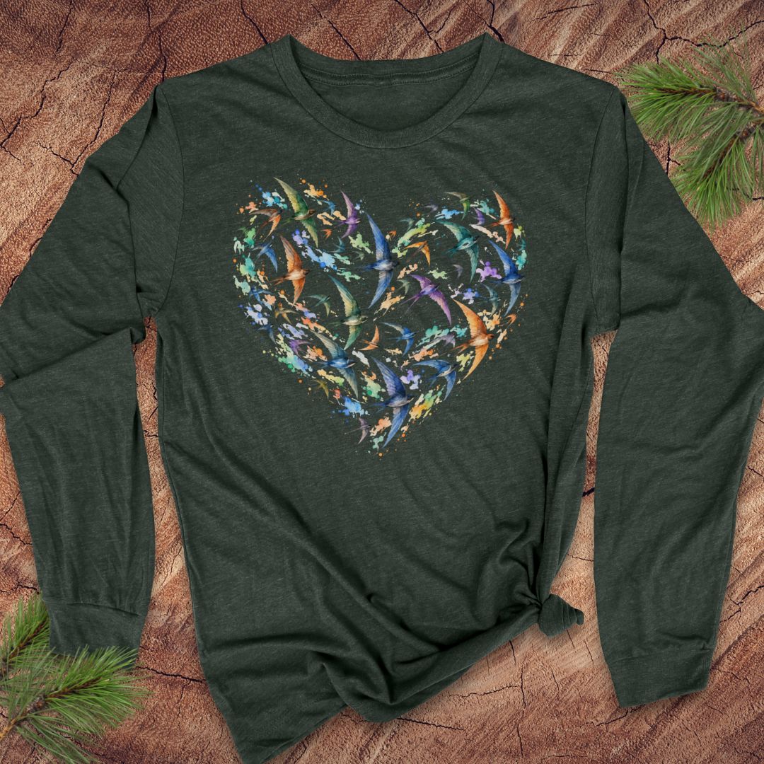 A dark forest green long-sleeve t-shirt with a colorful heart-shaped bird graphic made of small flying reptiles, laid flat on a rustic wood background with pine branches.