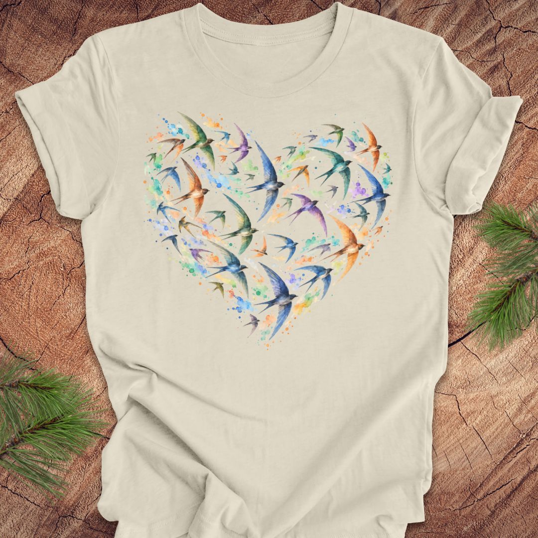 An off-white short-sleeve t-shirt laid flat on a rustic wood background, featuring a heart-shaped graphic made of colorful flying birds in shades of blue, green, purple, and orange.