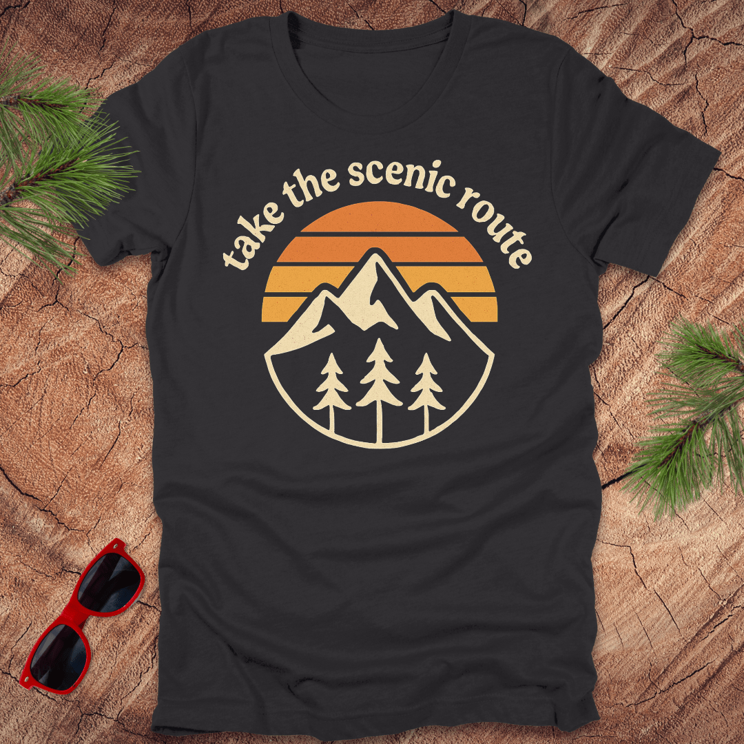 Black t-shirt with mountain and sunset design on a wooden surface with sunglasses and pine branches.