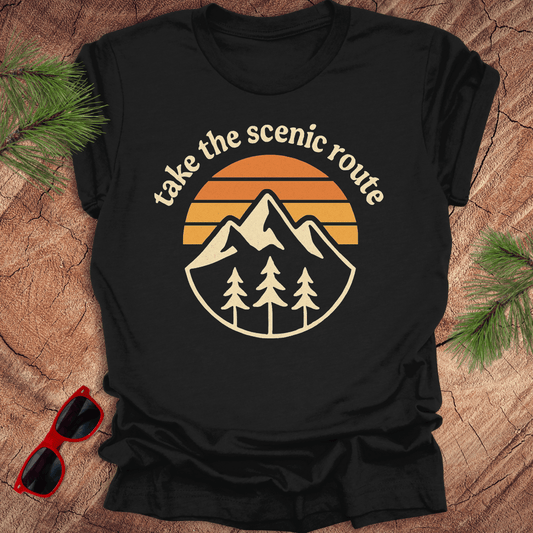 Black t-shirt with mountain and sunset design on a wooden surface with sunglasses and pine branches.