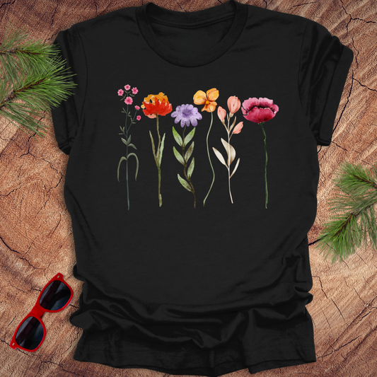 A black t-shirt with a floral design featuring various colors of tall flowers, displayed on a wooden surface with green leaves and red sunglasses to the side.