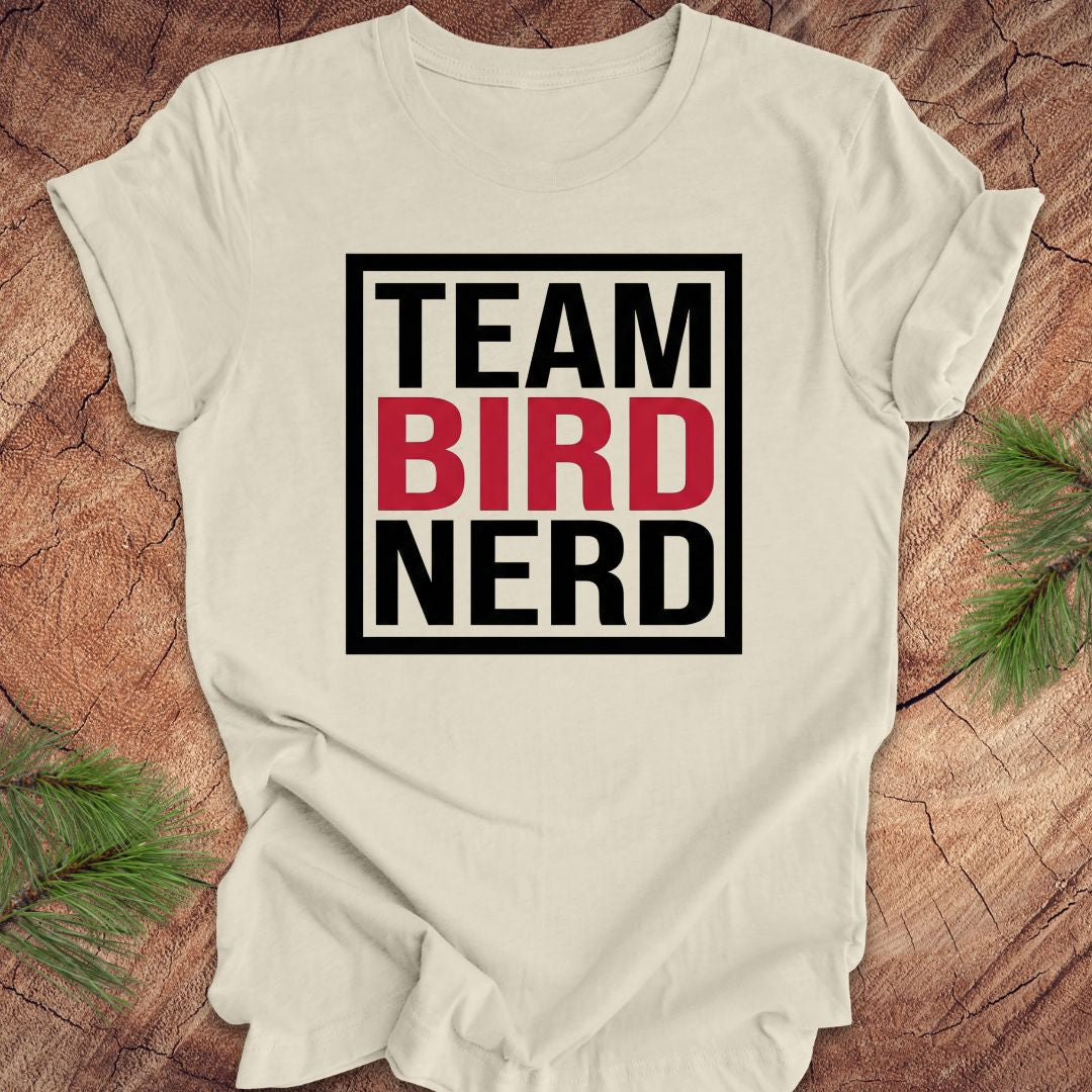 Beige t-shirt with 'Team Bird Nerd' text on a wooden surface with greenery.