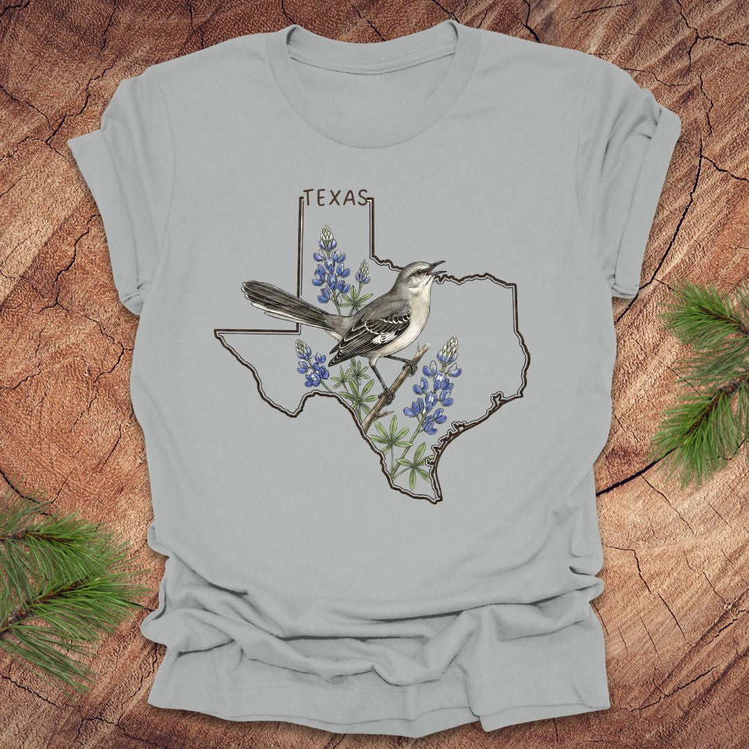 Silver t-shirt with Texas state outline with state bird, mockingbird and state flower bluebonnet design on a wooden surface