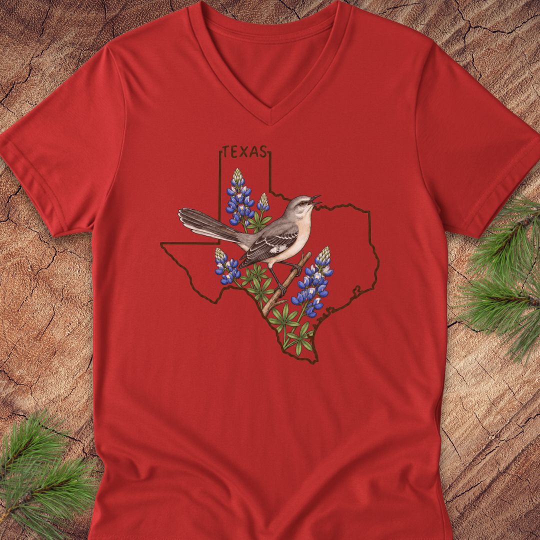 A red V-neck t-shirt laid flat on a rustic wood background with pine branches in the corners. The shirt features a graphic of the state outline of Texas with bluebonnets and a gray-and-white bird in the center, with the word 'TEXAS' above.
